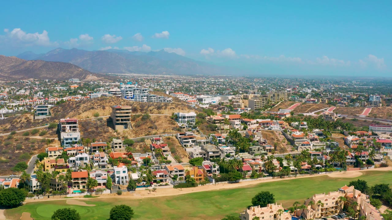 Los Cabos aerial view with white and terracotta-roofed homes below, tan hillside ridges through the middle, a dense city area beyond, and no visible people.