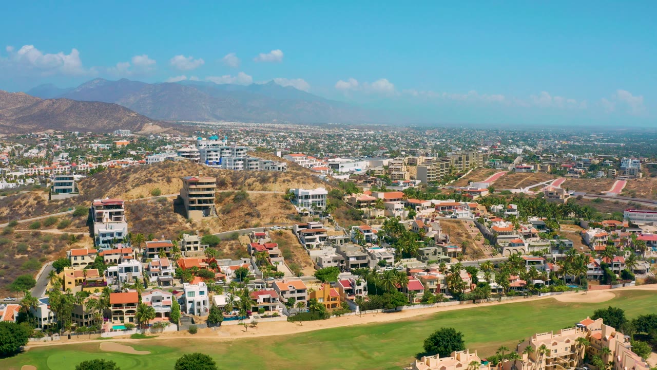 Aerial view across homes, hills, roads, green turf, and hazy mountains.