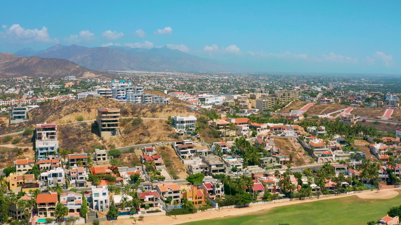 Opening aerial frame over Los Cabos, Mexico, with modern hillside homes, dry tan terrain, a green field edge, distant urban sprawl, hazy mountains, and no visible people.