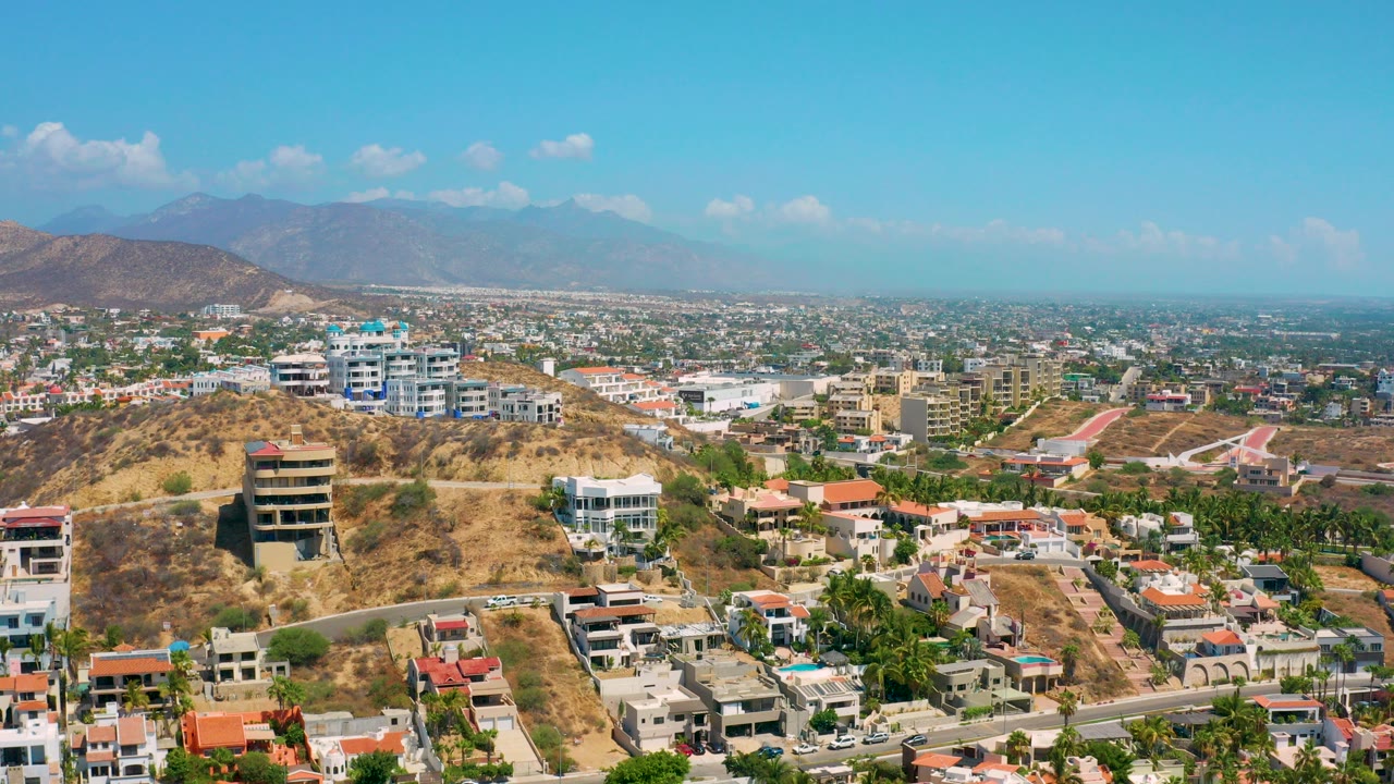 Aerial view over Los Cabos, Mexico with dry hillside homes in the foreground, a broad suburban area beyond, distant mountains, blue sky, and no visible people.