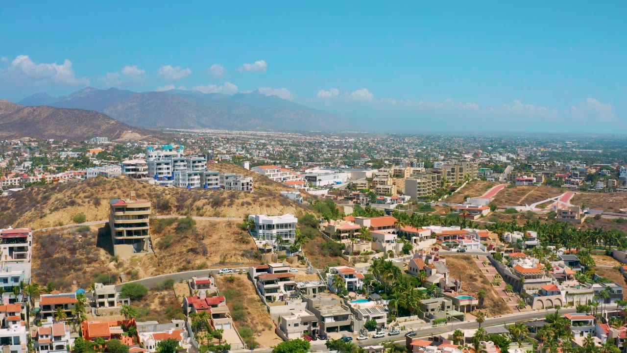 Drone perspective over Los Cabos, Mexico showing modern houses on dry slopes, a road crossing the lower frame, dense low-rise development, hazy mountains, and no visible people.