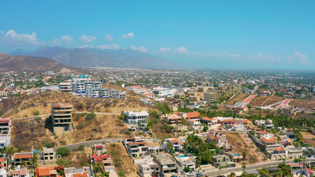 Wide aerial frame in Los Cabos, Mexico with a tan hillside, multi-story homes, paved roads, scattered greenery, distant urban development, mountains, and no visible people.