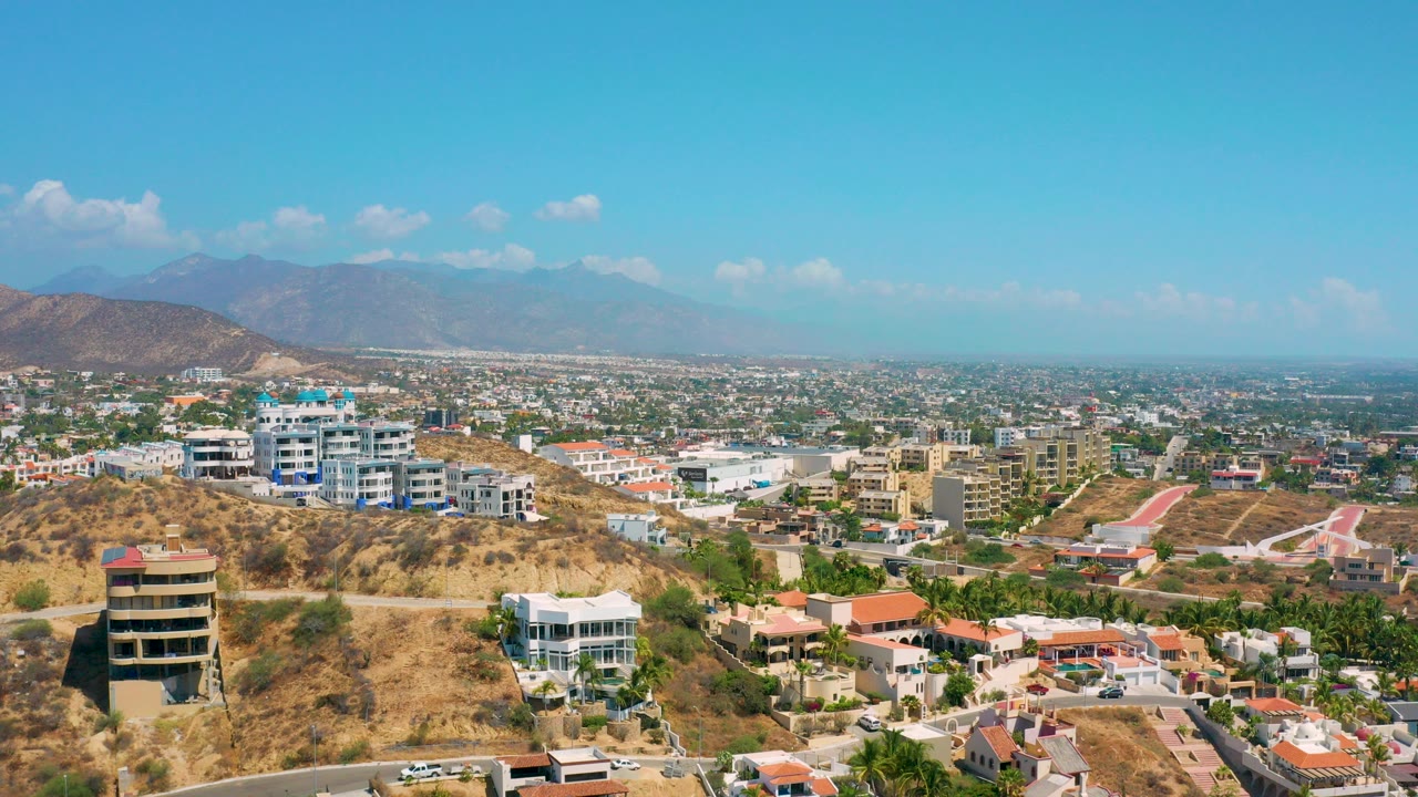 Opening aerial frame of Los Cabos, Mexico with dry hills in the foreground, white residential buildings, flat urban land, hazy mountains, blue sky, and no visible people.