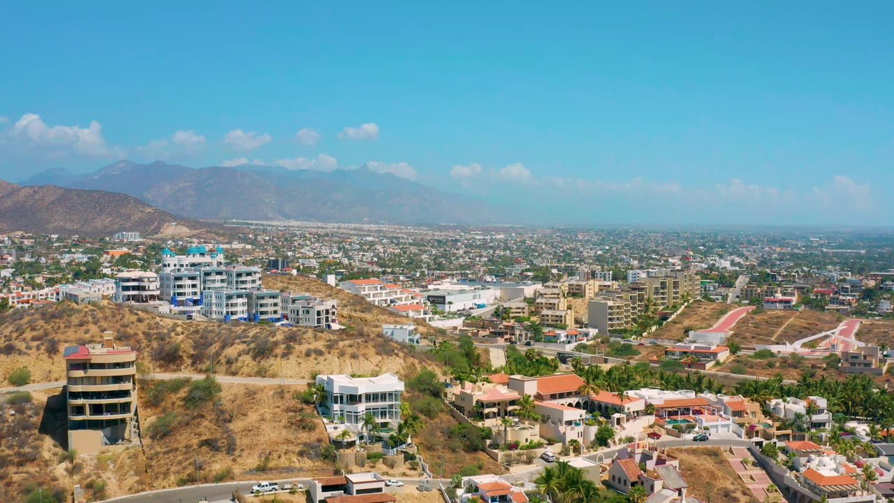 Los Cabos aerial view with a tan hillside, clustered white and blue buildings, curving roads, low city blocks, distant mountain ridges, and no visible people.