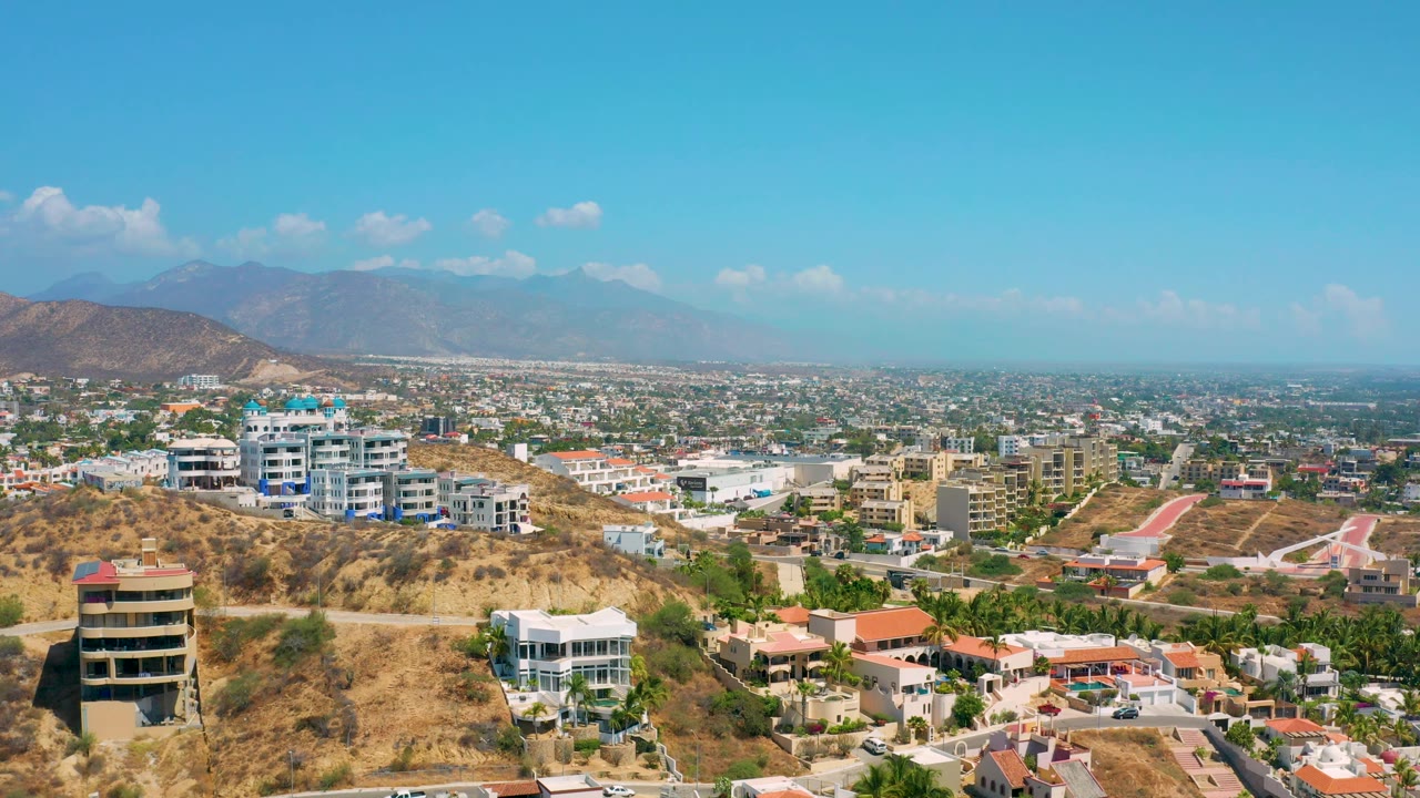 Los Cabos frame showing residential roofs below a dry ridge, a rounded dark building near the lower left, broad urban development, distant mountains, and no visible people.