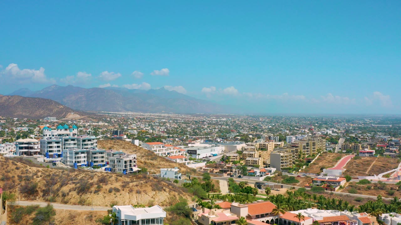 Opening frame of Los Cabos, Mexico from above a dry hillside, with multi-story homes in the foreground, a broad city valley beyond, distant mountains, scattered clouds, and no visible people.