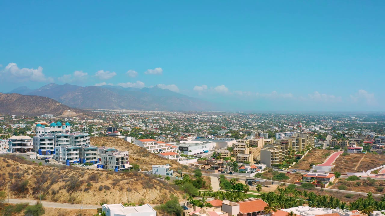 Bright Los Cabos overview with white and blue multi-story buildings on a dry hillside, tan undeveloped slopes, a spread of city structures, hazy mountains, and no visible people.