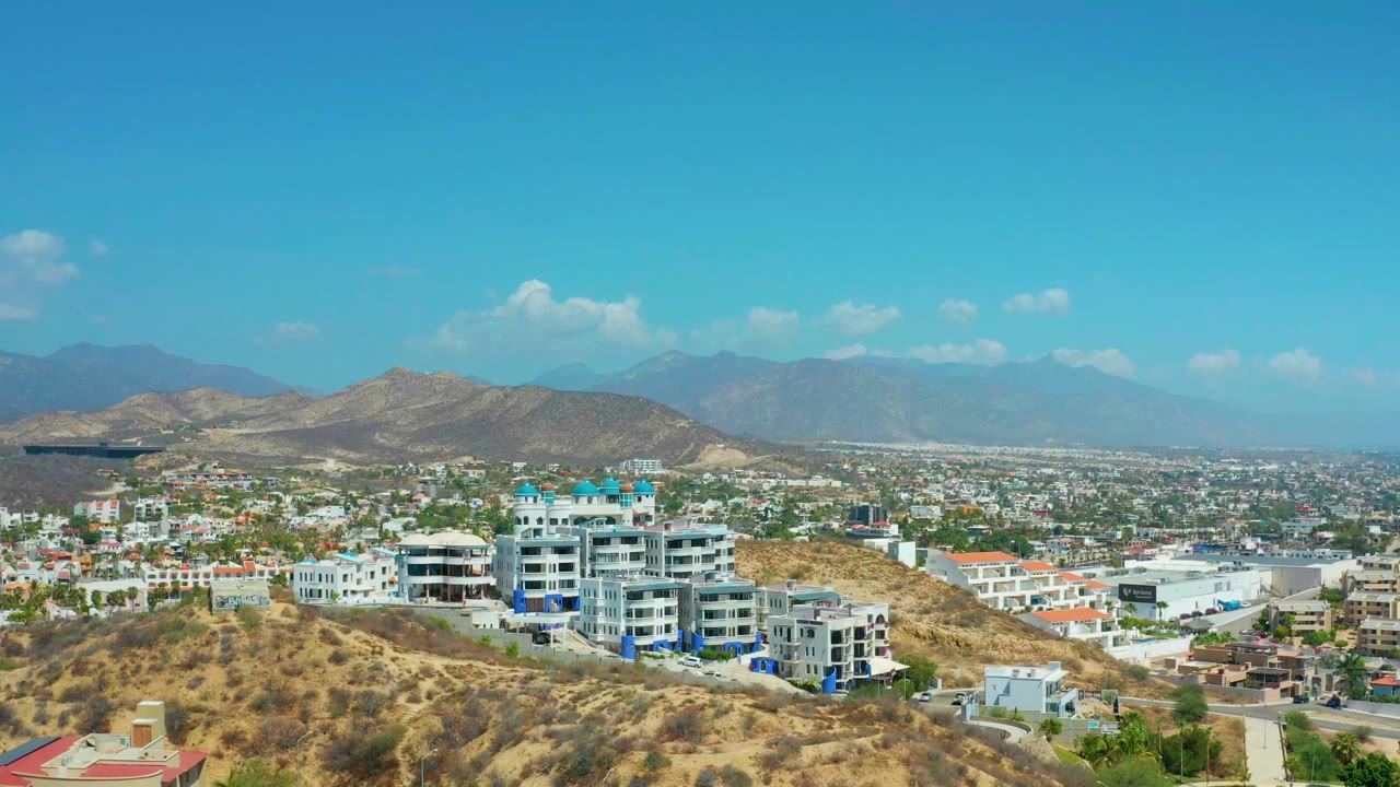 Opening frame of Los Cabos, Mexico from a high vantage, with dry hills in front, white resort buildings below the ridge, distant mountains, bright blue sky, and no visible people.