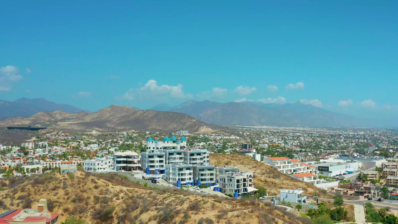 Los Cabos, Mexico hillside resort view with the main white buildings near center, scrub-covered slopes in the foreground, a broad townscape behind, hazy mountains, and no visible people.