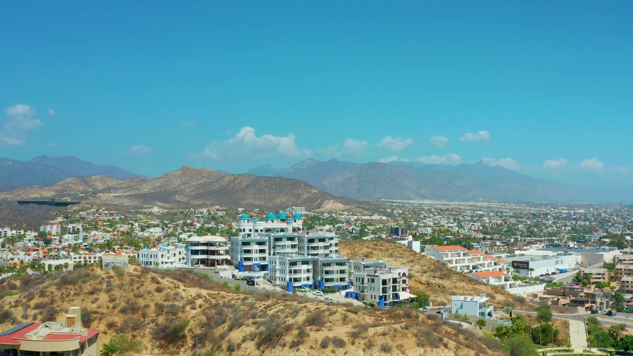 Wide Los Cabos, Mexico view with resort buildings slightly right of center, tan hills across the foreground, urban blocks across the valley, soft mountain silhouettes, and no visible people.