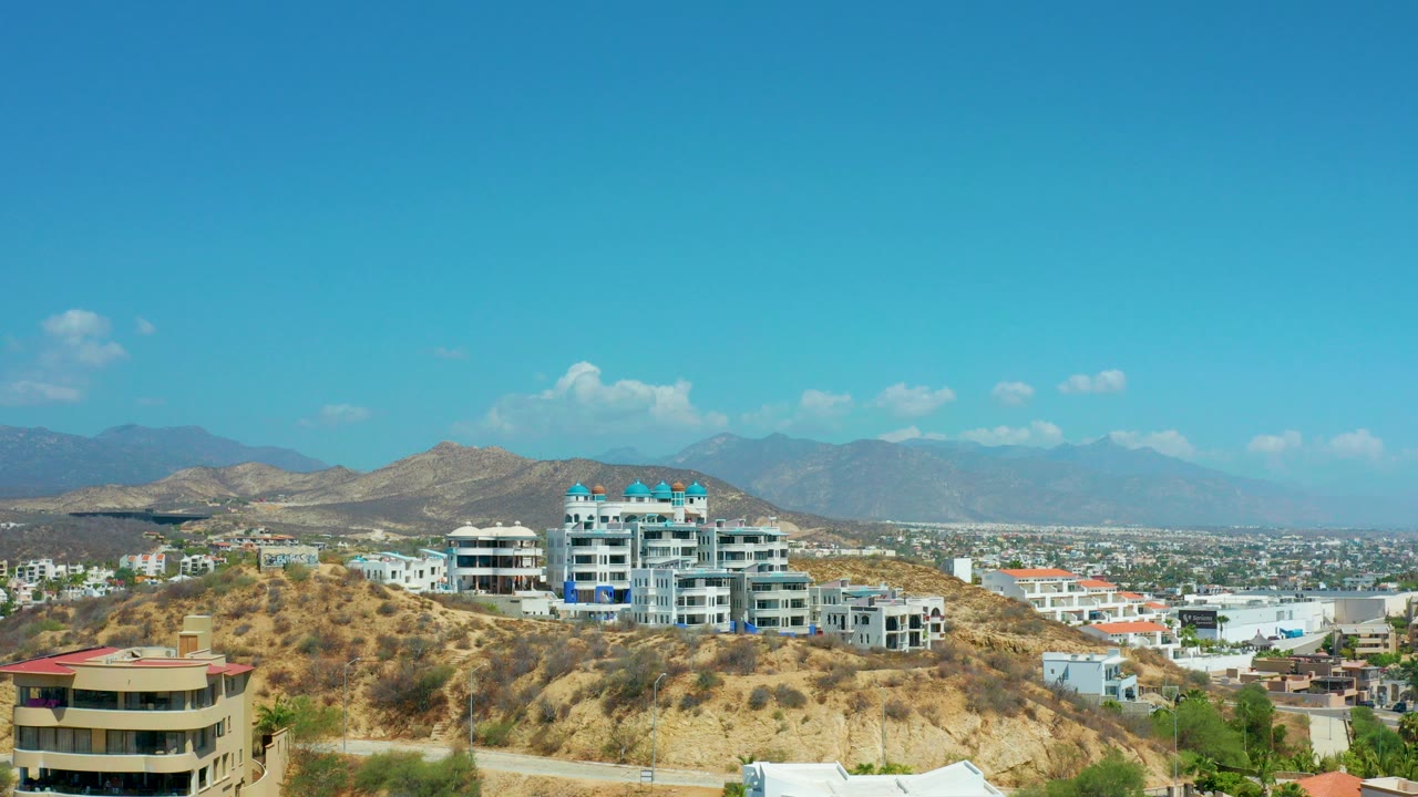 Opening frame of Los Cabos, Mexico, showing white and beige hillside buildings, dry tan slopes, a distant city basin, blue mountain ridges, and no visible people.