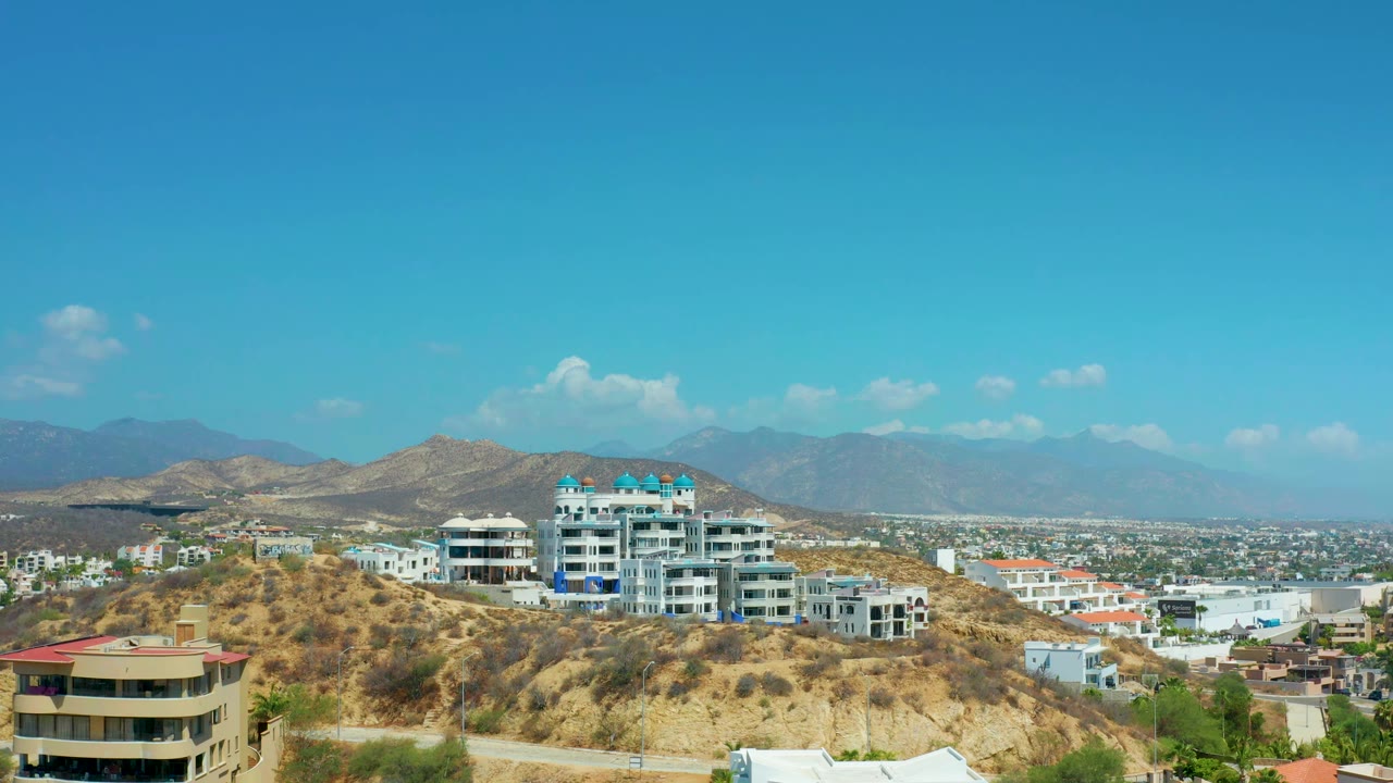 Early frame in Los Cabos, Mexico, with modern white buildings centered on the ridge, a beige foreground building at lower left, distant urban blocks, mountains, and no visible people.
