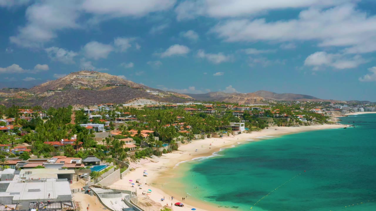 Opening aerial frame over Palmilla Beach Cabo, Mexico, with white-roofed beachfront buildings at lower left, a pale sand curve, turquoise water, and dry mountains under scattered clouds.