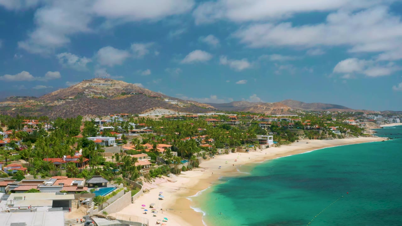 Aerial view at 0:01 over Palmilla Beach Cabo, Mexico, showing the shoreline slightly farther along the beach, with turquoise water on the right, palm-covered buildings inland, hills in back, and no visible people.