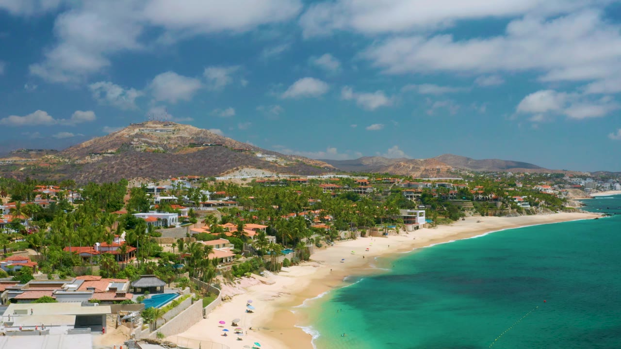 At 0:02, the Palmilla Beach Cabo aerial view shows more open water to the right, a light sand beach below the palms, hillside terrain across the background, and no visible people.