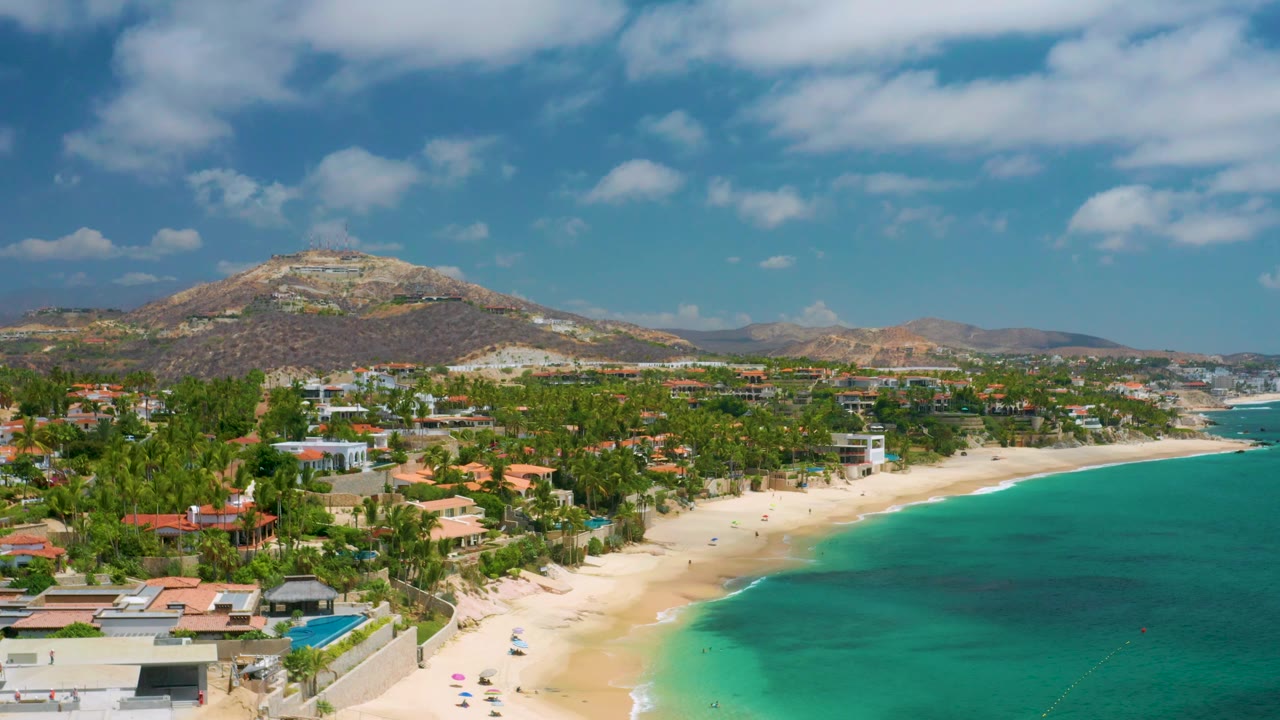 Aerial frame of beach houses, palms, pale sand, turquoise water, and rocky hills.