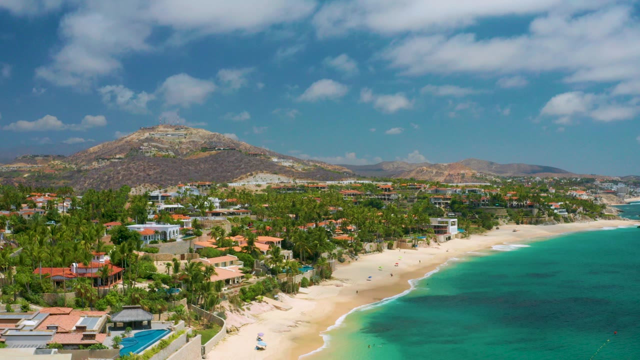 Opening aerial frame over Palmilla Beach Cabo, Mexico, with turquoise water at right, pale sand below, palm-lined homes at left, hills behind, and no visible people.
