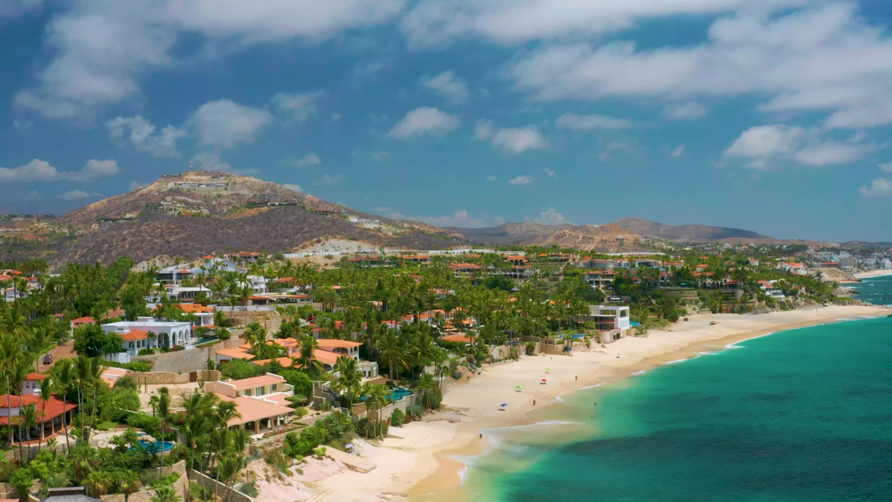 Wide aerial shoreline with beach homes, palms, blue water, and hills.