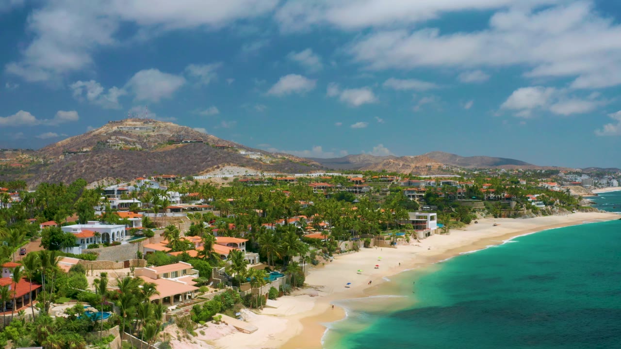 Aerial frame over Palmilla Beach Cabo, Mexico, with the sandy beach in the lower center, turquoise bay at right, palm-covered properties at left, hills behind, and no visible people.