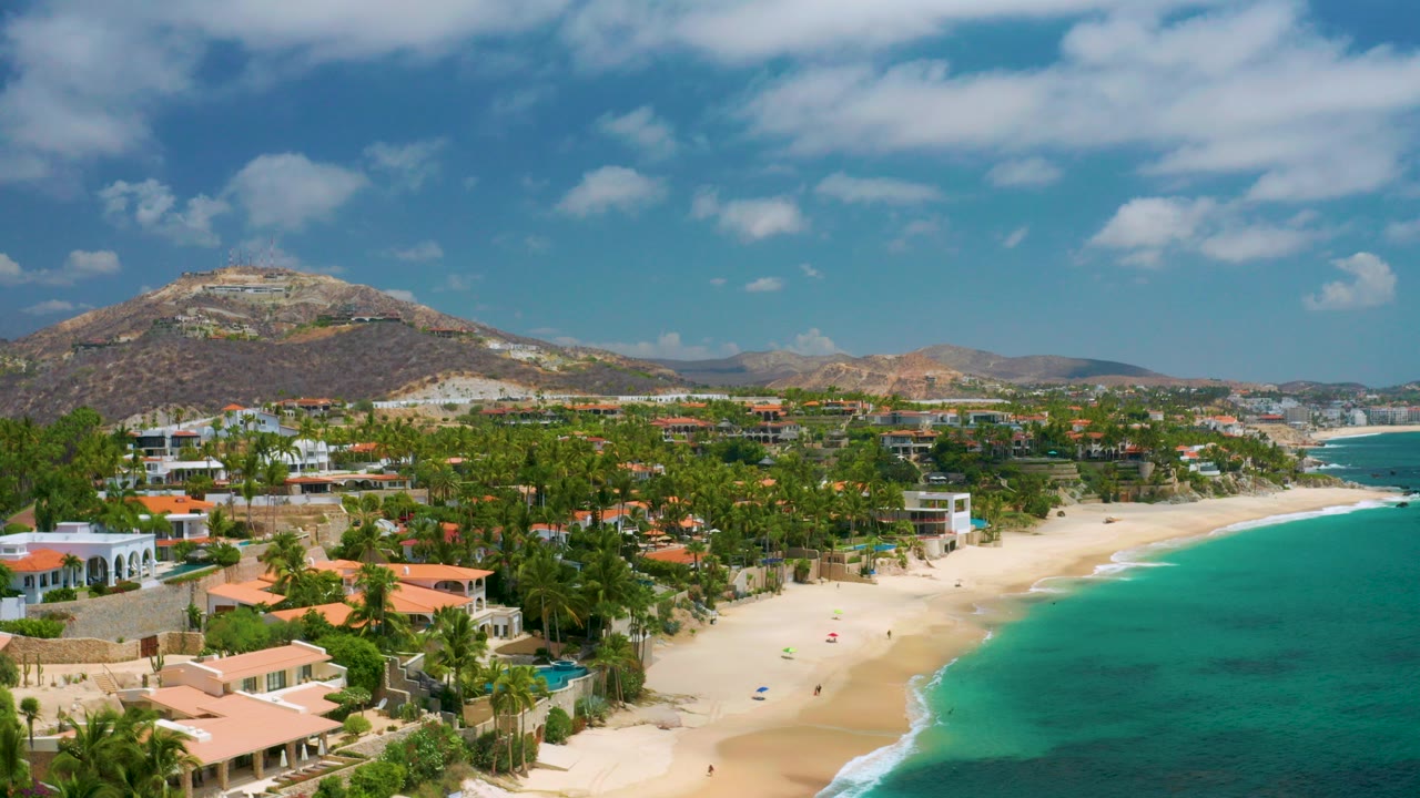 Aerial view over Palmilla Beach Cabo, Mexico with beach houses and palms in the foreground, white sand and turquoise water to the right, dry hills behind, and no visible people.
