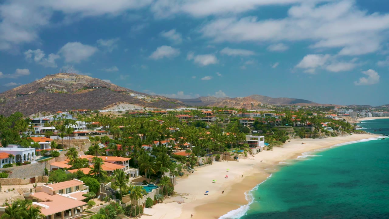 Palmilla Beach Cabo, Mexico appears from above with beachfront homes at left, a broad strip of light sand below, blue-green water at right, hills in back, and no visible people.