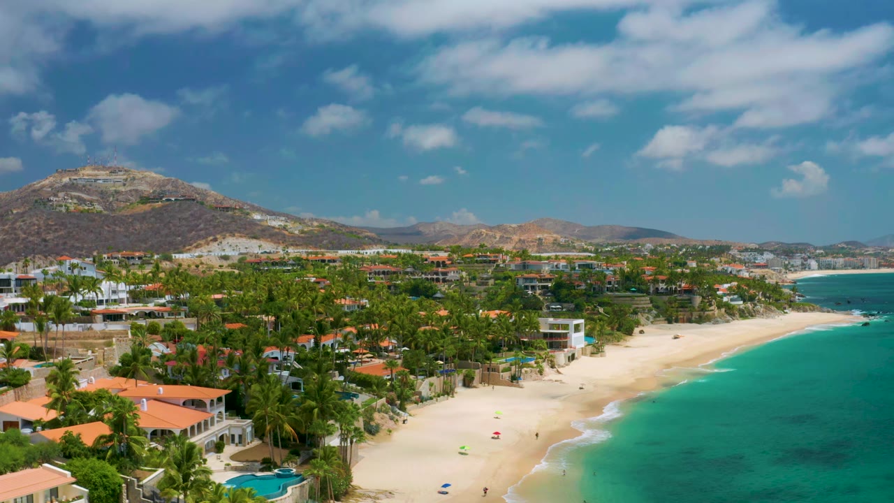 Wide aerial coastline with houses, palms, beach, and blue-green water.