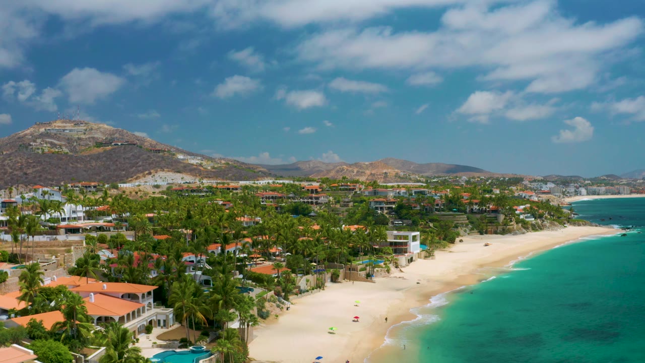 Aerial view in Palmilla Beach Cabo with more ocean visible on the right, beachfront villas and palms along the sand, low mountains in the distance, and no visible people.