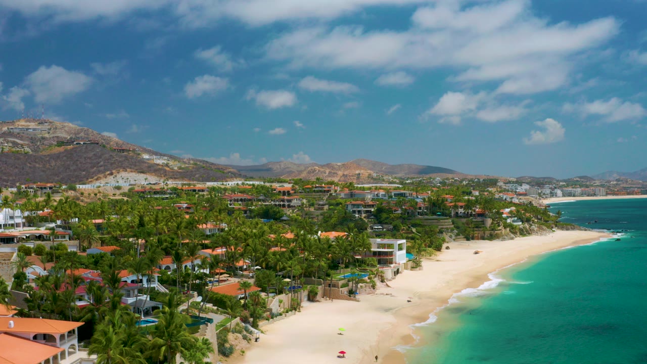 Opening frame over Palmilla Beach Cabo, Mexico, with turquoise water beside pale sand, palm-covered villas, dry hills in the distance, scattered clouds, and no visible people.