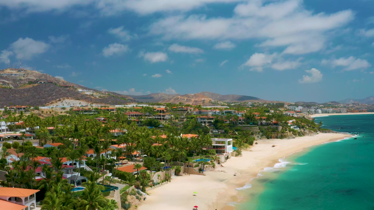 A wide Palmilla Beach Cabo, Mexico frame shows the beach running below the palms, teal water along the right edge, coastal villas and pools inland, hills on the horizon, and no visible people.