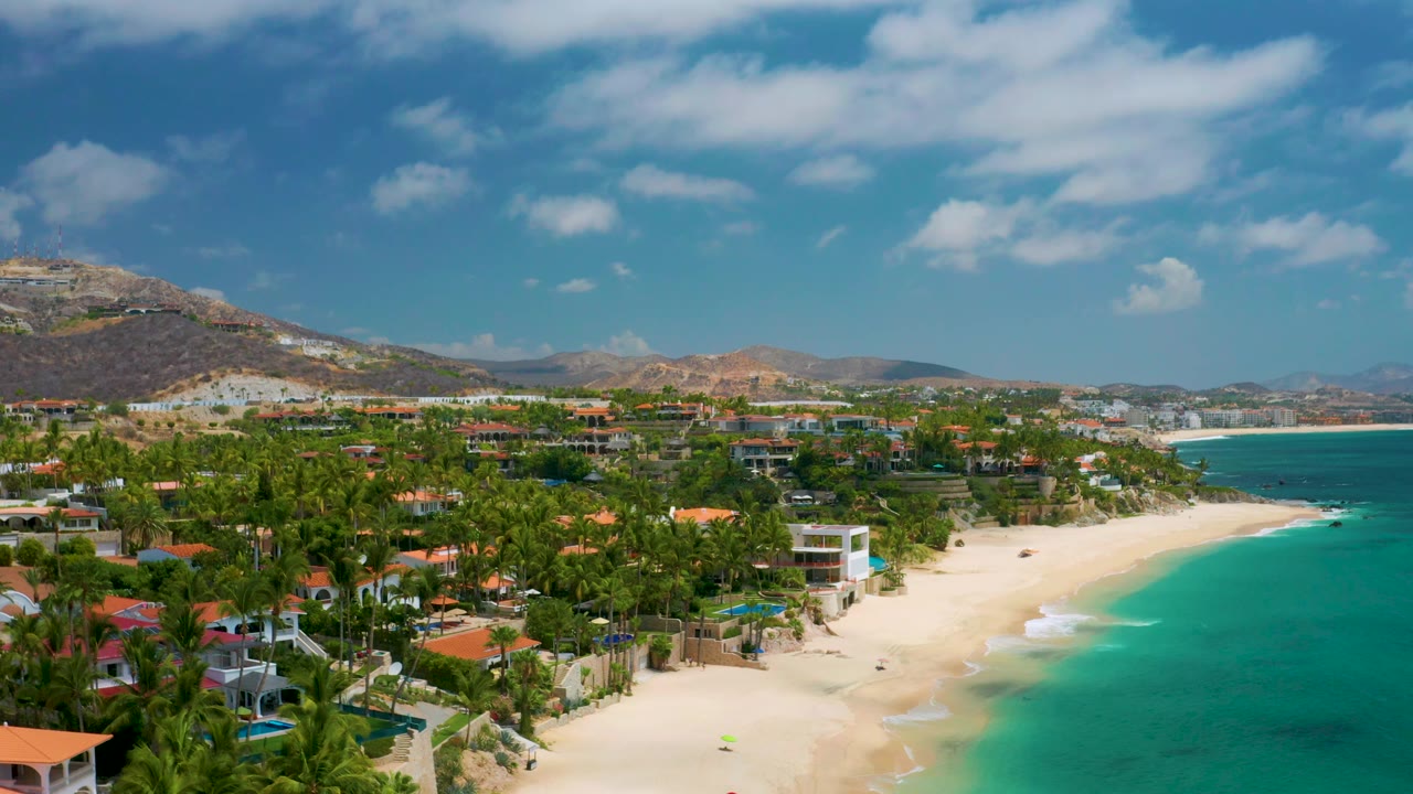 Elevated beach view with palms, villas, surf line, sea, and hills.