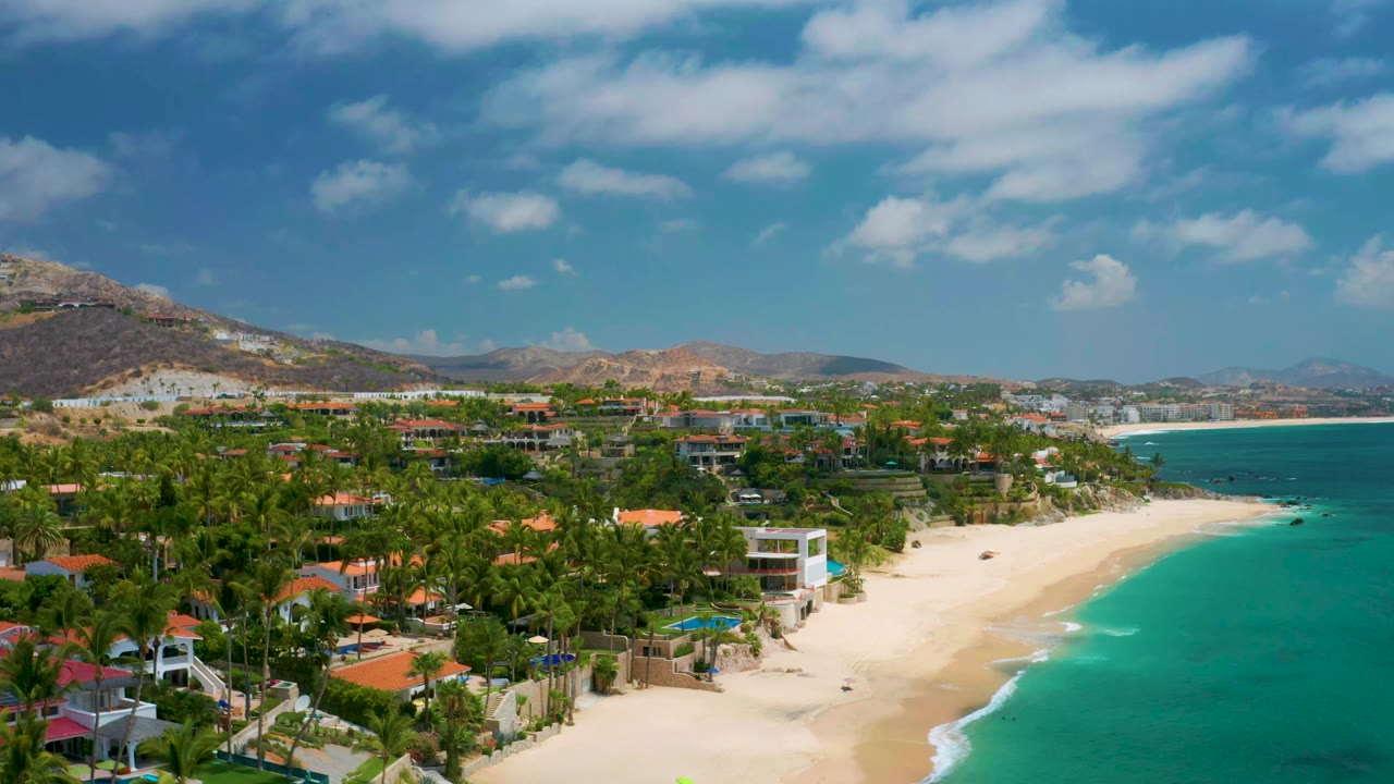 Wide coastal aerial with palms, beach houses, bright sand, and blue-green water.