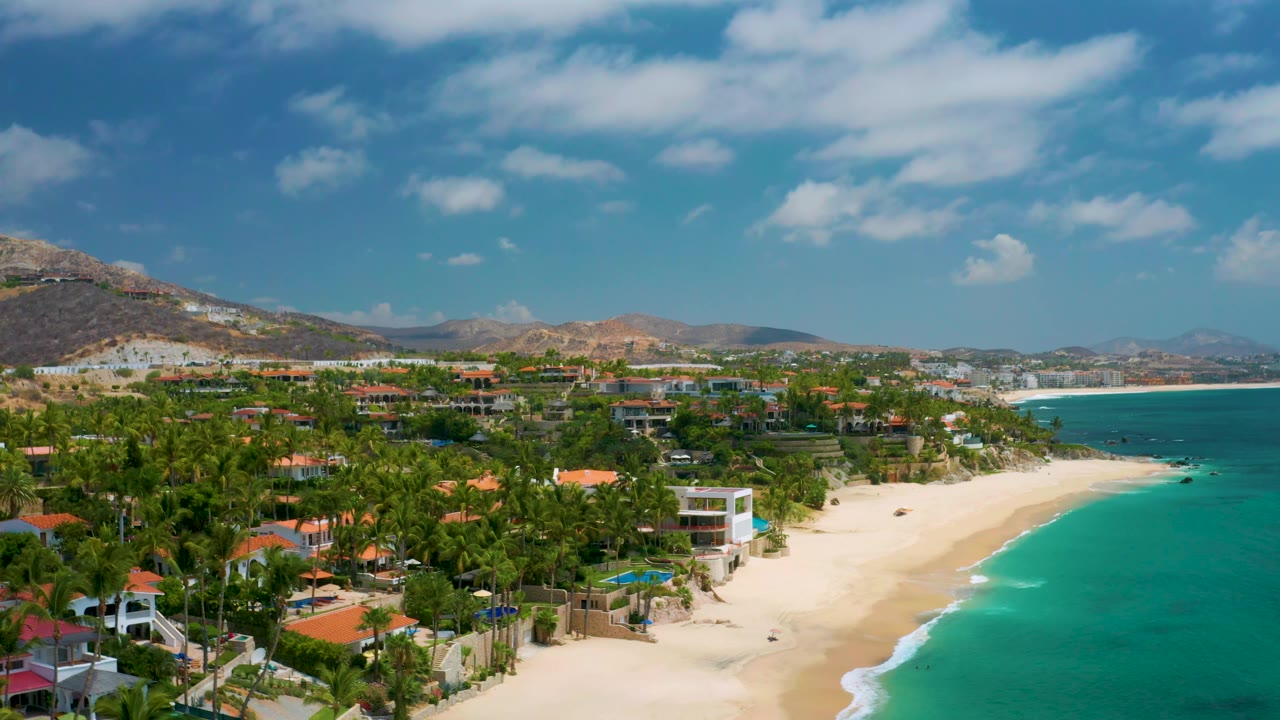 Aerial frame from Palmilla Beach Cabo, Mexico, showing the surf line farther along the sandy beach, dense palms, low coastal buildings, distant shoreline, and no visible people.