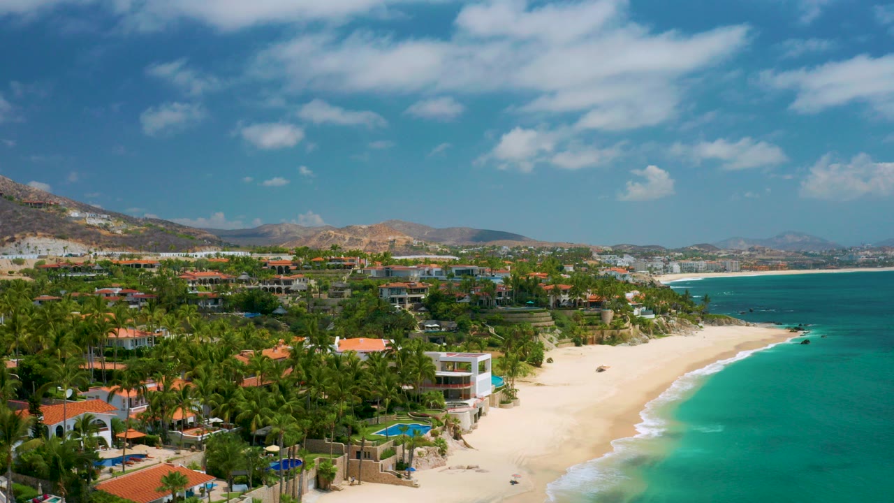 Wide aerial frame of Palmilla Beach Cabo, Mexico with a sandy beach, turquoise sea, palms, beachfront houses, distant hills, blue sky, and no visible people.