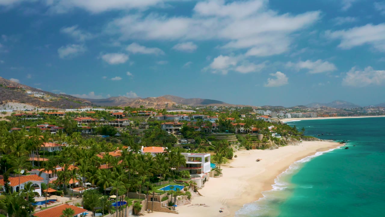 Wide Palmilla Beach Cabo, Mexico aerial with red-roofed houses among palms, a bright sand beach, turquoise bay water, low surf, distant hillside buildings, and no visible people.