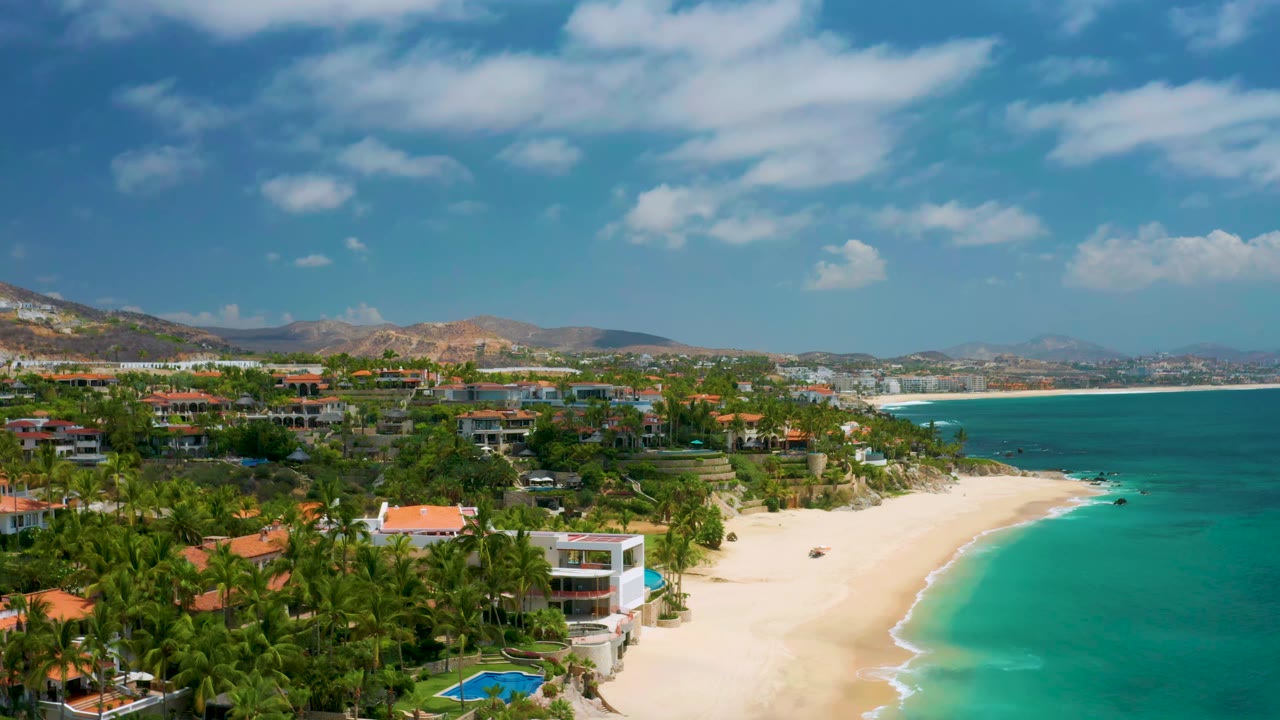 Palmilla Beach Cabo aerial view with resort roofs and dense palms in the foreground, a curving sand beach at center, blue-green sea to the right, and no visible people.