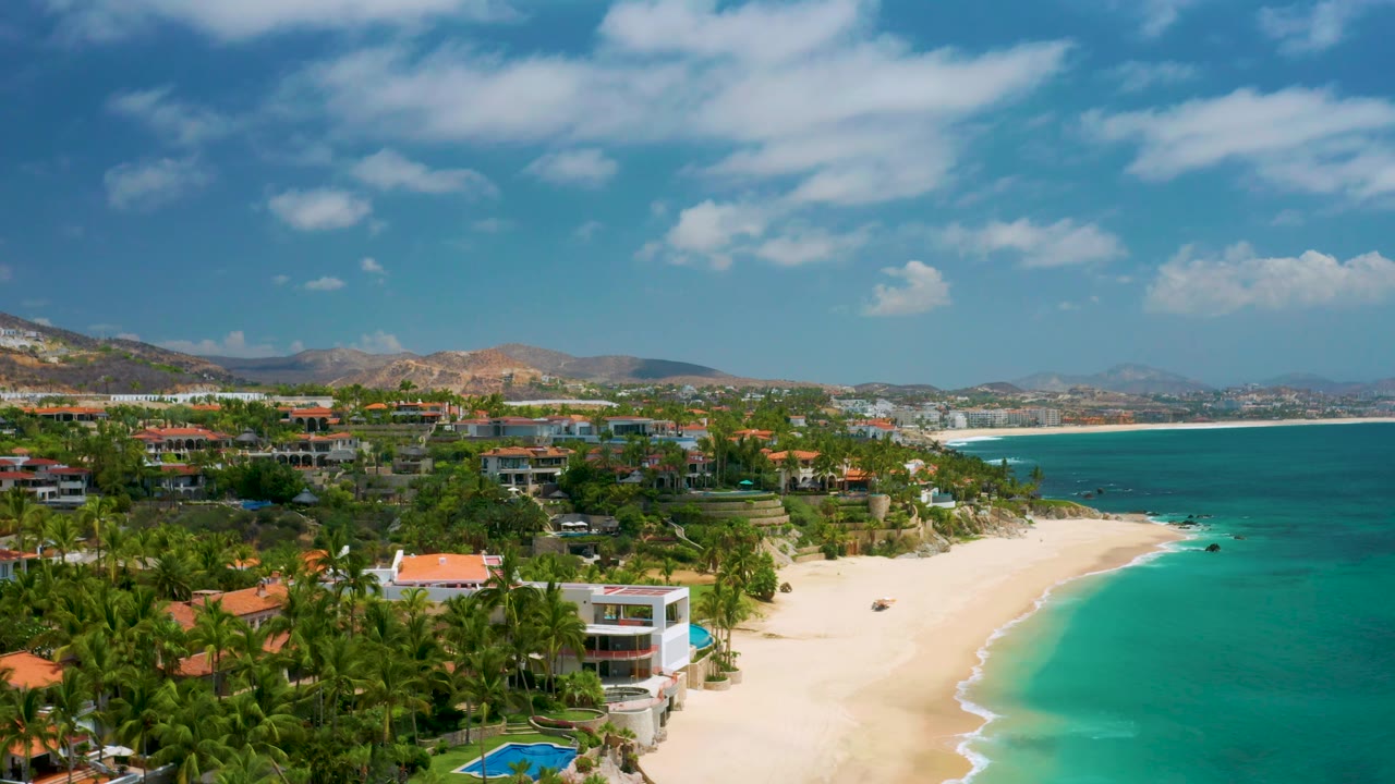 Aerial frame over Palmilla Beach Cabo showing a white beachfront structure below the palms, red-roofed buildings inland, a bright sand strip, turquoise water, and no visible people.