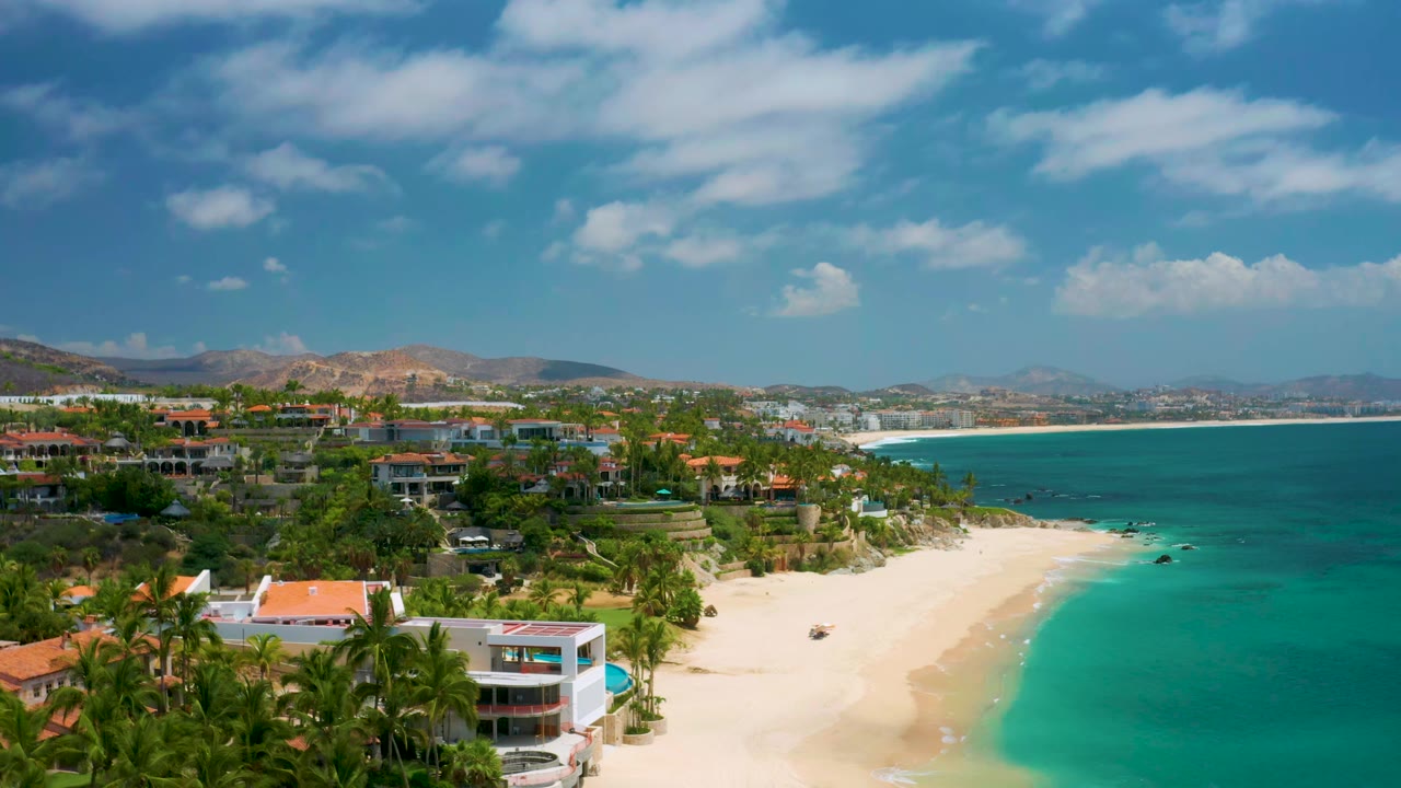 Palmilla Beach Cabo aerial view one second in, showing the sandy shoreline centered below the bay, palms and low buildings in the foreground, mountains beyond town, and no visible people.