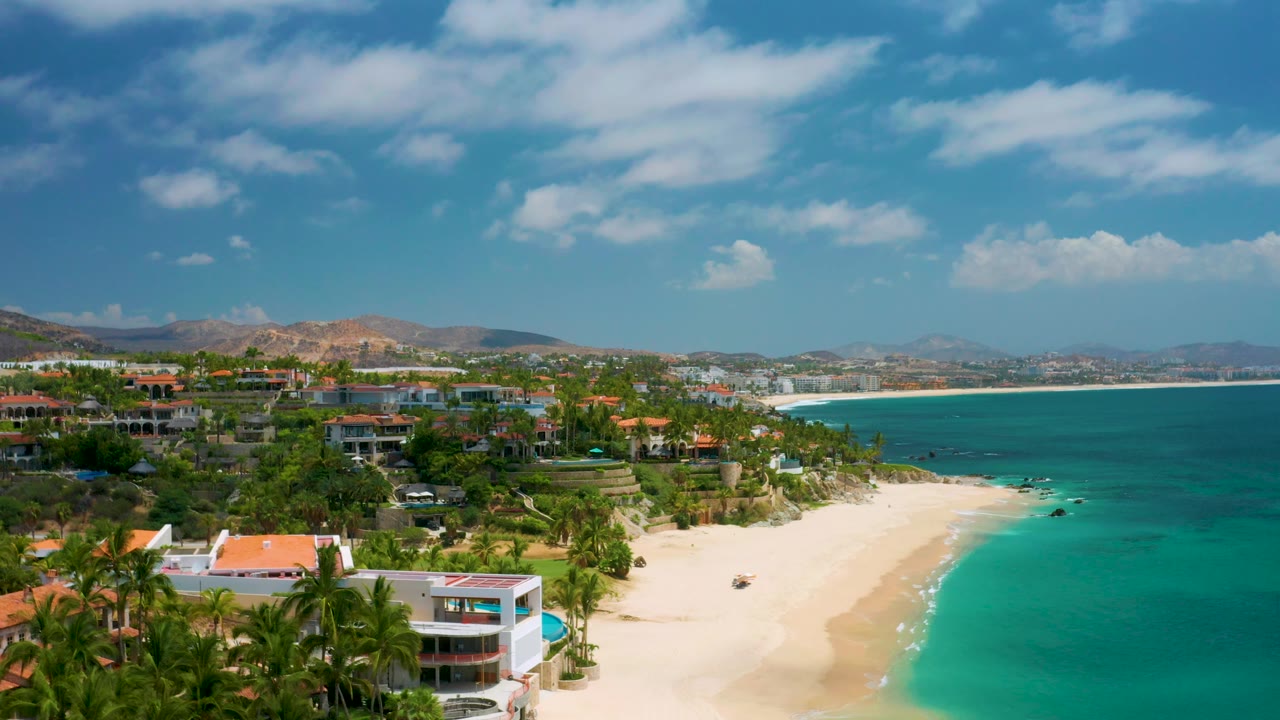 Aerial frame at Palmilla Beach Cabo with a white building and palms near the lower edge, empty sand along the waterline, turquoise sea to the right, and no visible people.