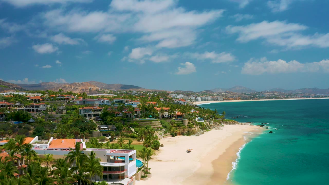 Aerial view along sandy beach beside calm water and resort-area greenery.