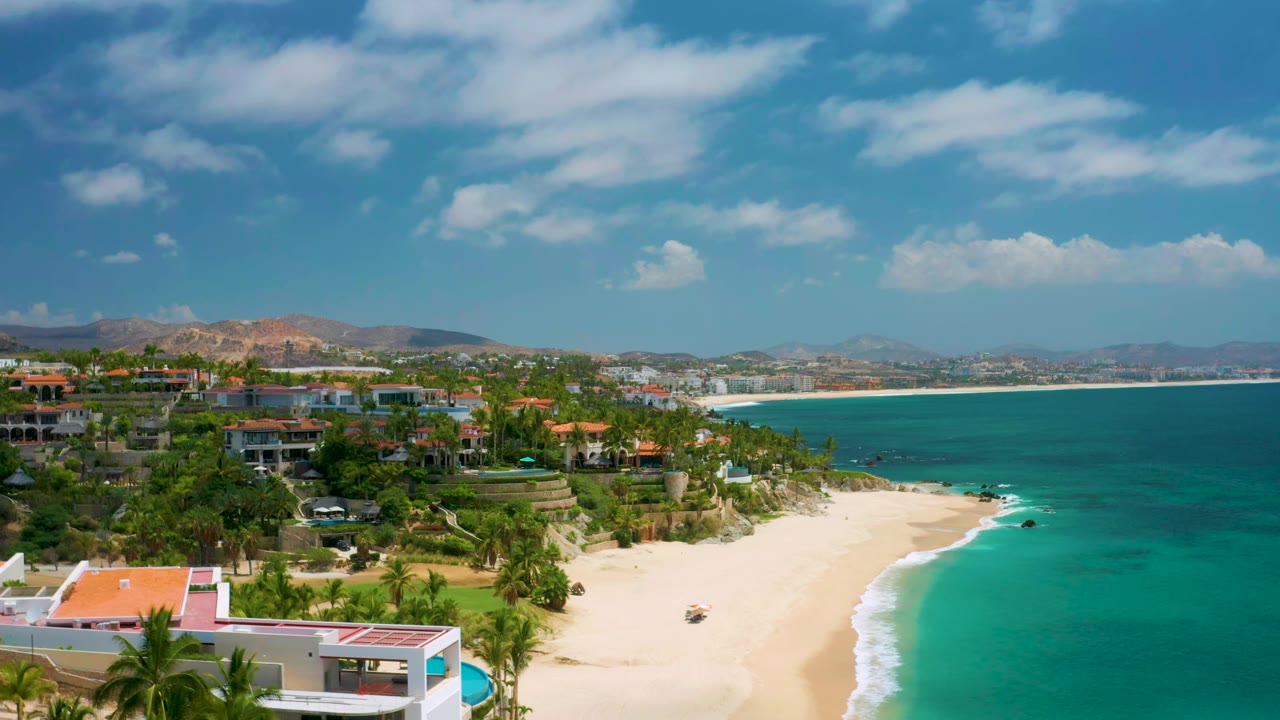 Wide aerial coastline view with sandy beach, teal water, palm-lined buildings, and hills.