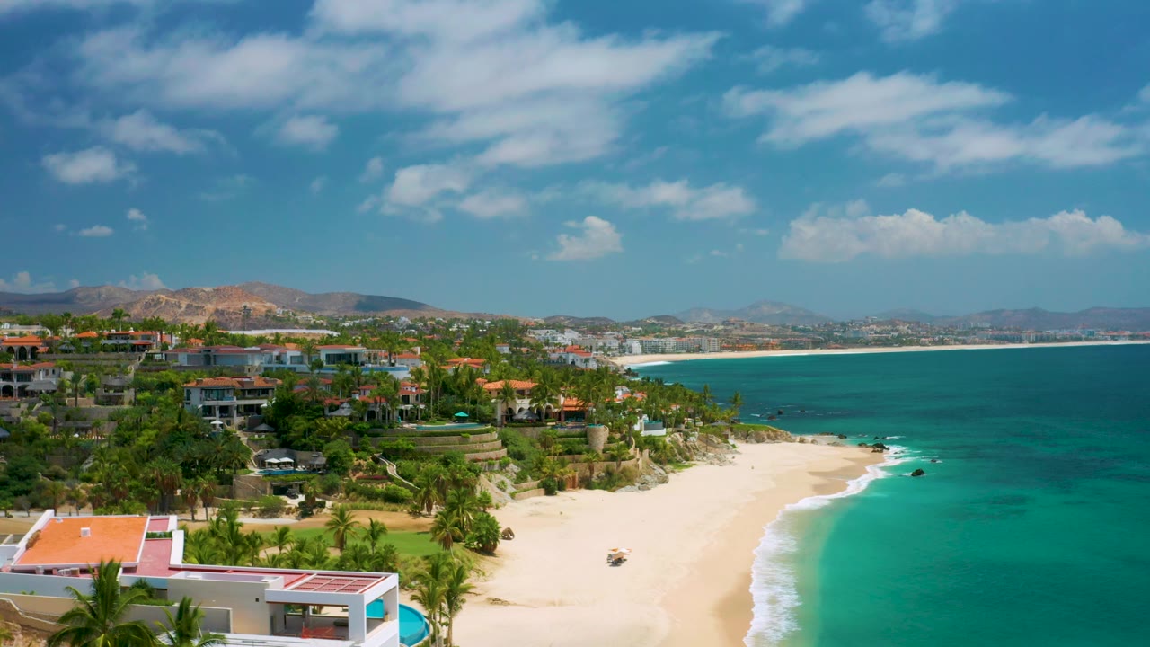 Frame from the drone-height view over Palmilla Beach Cabo, Mexico, with red-roofed buildings low in the foreground, palms above the sand, bright turquoise water, hills, and no visible people.