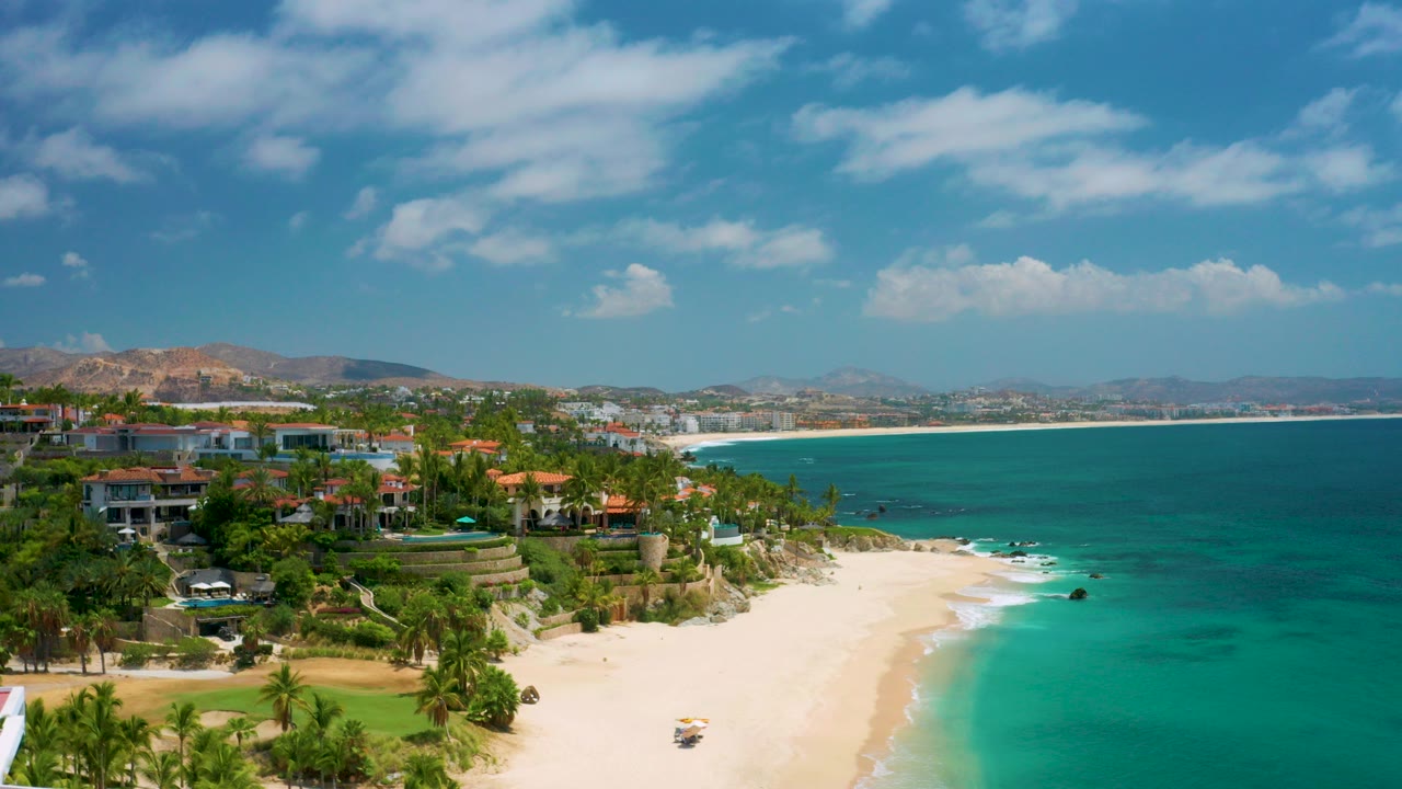 Opening aerial view at Palmilla Beach Cabo, Mexico, showing oceanfront homes above a pale sandy beach, turquoise water, distant hills, scattered clouds, and no visible people.