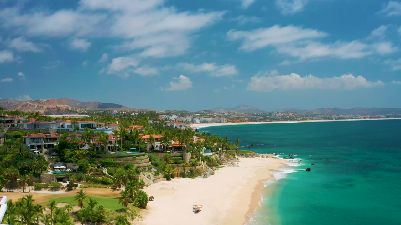 Wide coastline with beachfront homes, palms, pale sand, and blue-green water.
