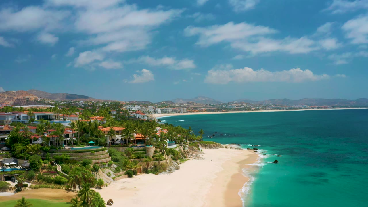 Palmilla Beach Cabo coastline with the residential hillside more centered, pale sand curving along the surf, deep blue water beyond, distant hills, and no visible people.