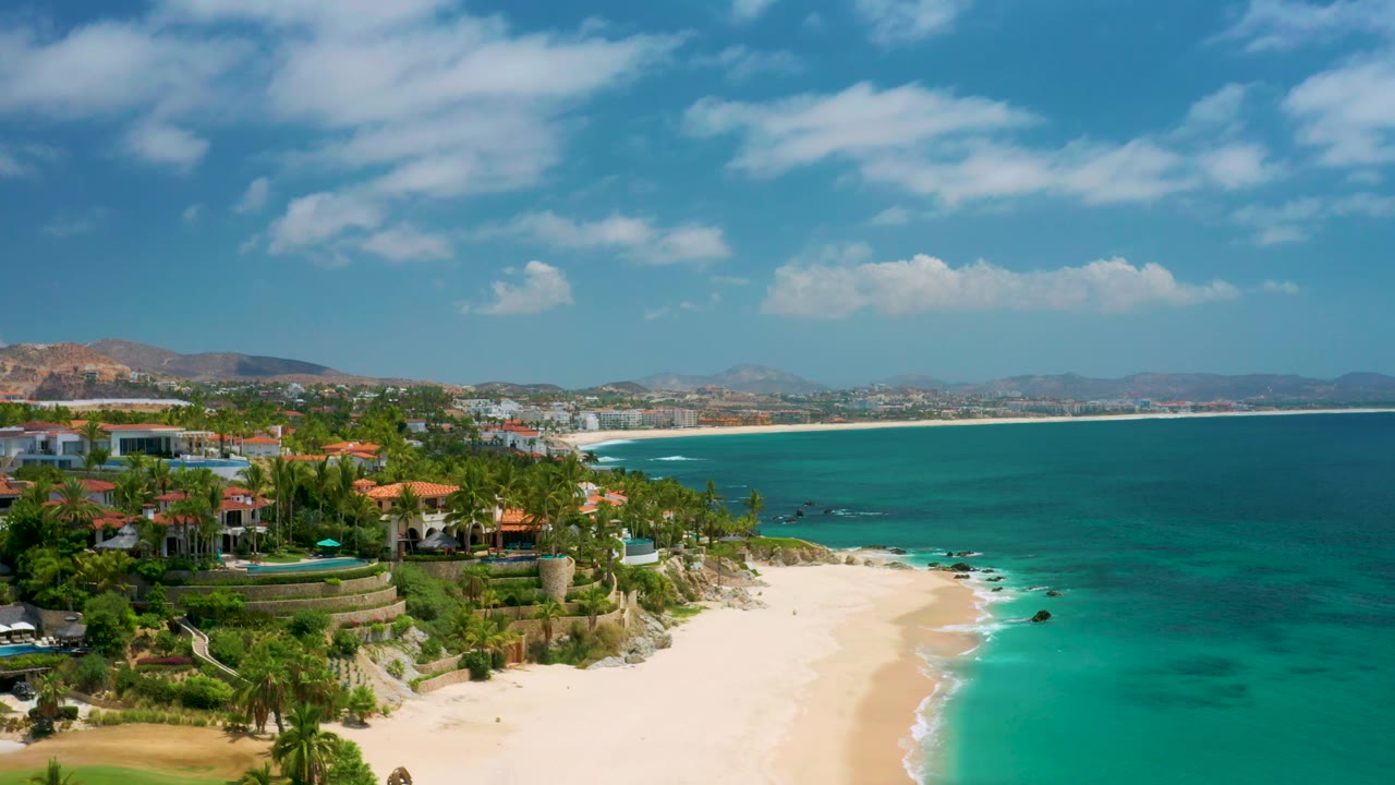 Elevated frame of Palmilla Beach Cabo with cliffside buildings and palms at left, white beach in the lower center, clear turquoise shallows, open bay water, and no visible people.