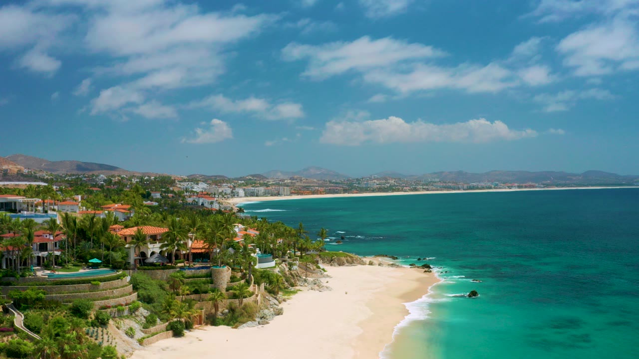 Opening aerial view of Palmilla Beach Cabo, Mexico, with red-roof villas and palms at left, pale sand below, turquoise water to the right, distant hills on the horizon, and no visible people.