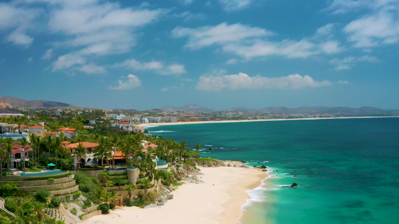 Aerial view over Palmilla Beach Cabo with the foreground villas slightly farther left, a broad band of turquoise sea on the right, white surf at the sand, and no visible people.