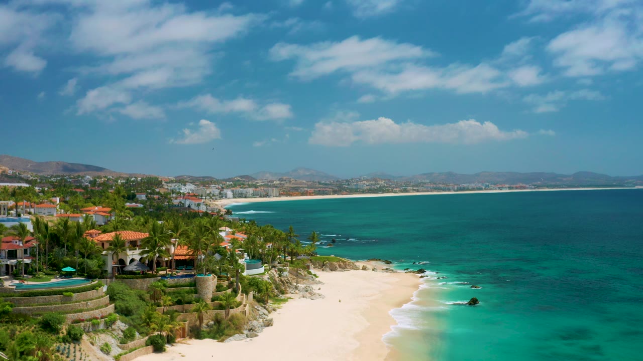 Aerial beach view with villas at left, wide ocean at right, and hazy hills beyond.