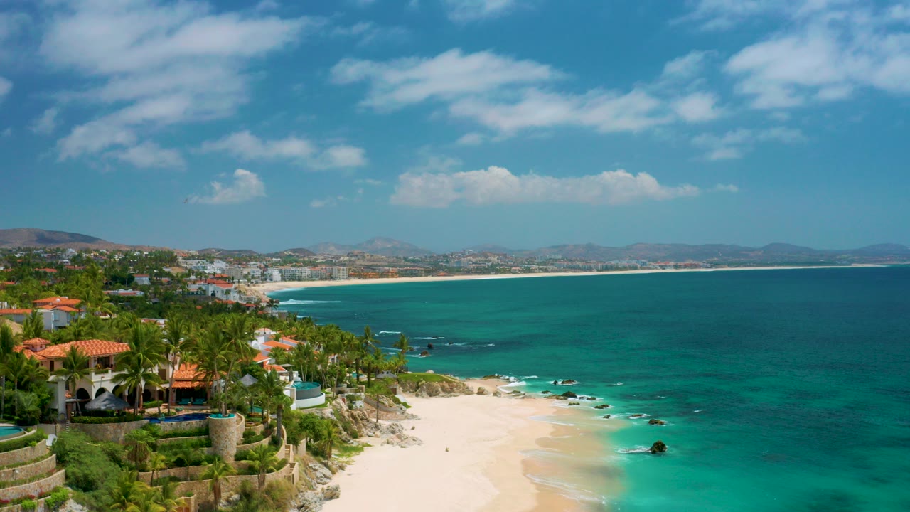 Aerial frame over Palmilla Beach Cabo, Mexico with red tile roof buildings and palms at left, turquoise water at right, pale beach below, and no visible people.
