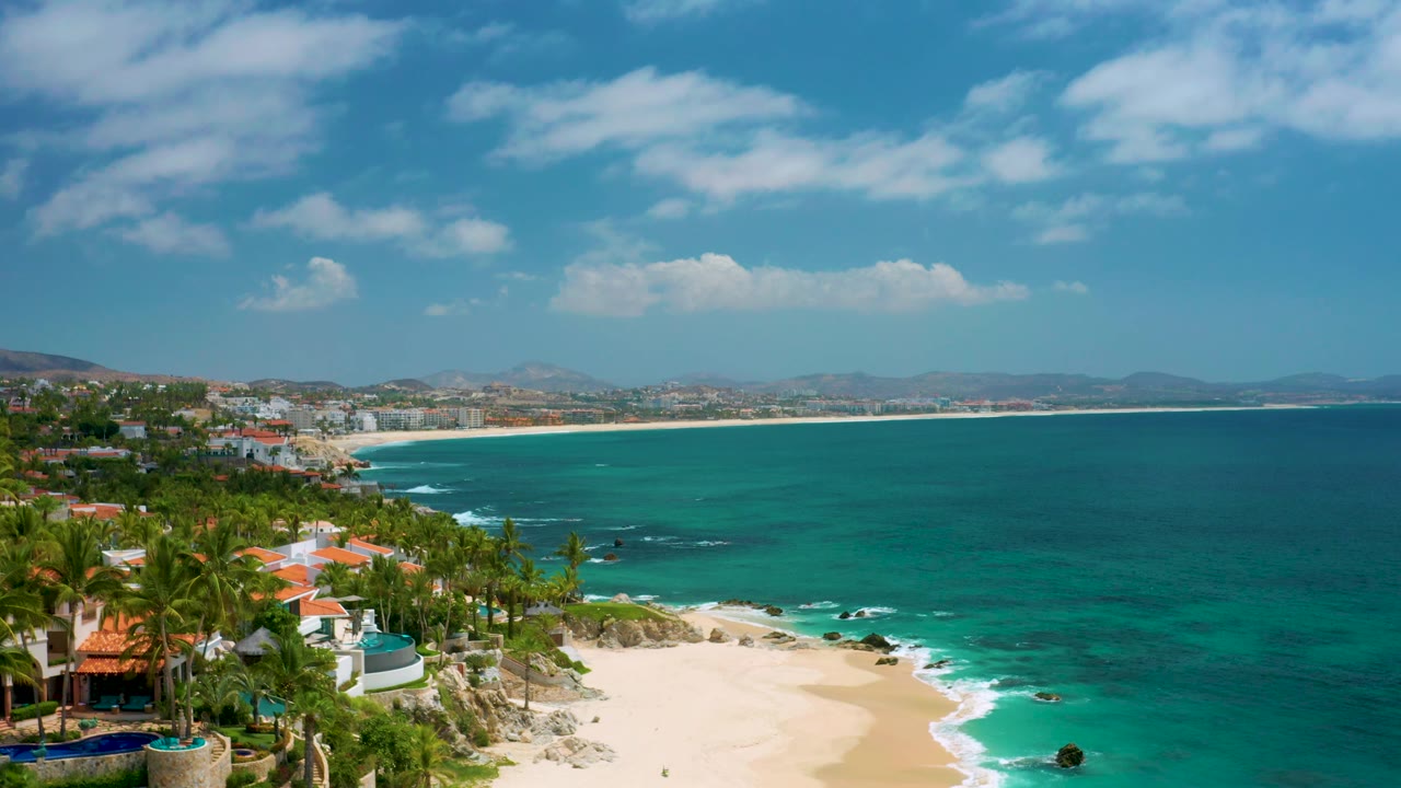 At 0:02, the aerial view shows Palmilla Beach Cabo with the beach cove lower left, stronger teal water near shore, homes and palms along the bluff, distant coast, and no visible people.